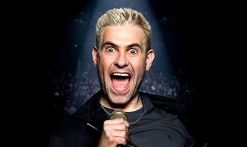 Simon Brodkin with his eyes and mouth very wide open staring down the camera under a spotlight holding a microphone