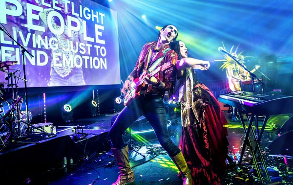 The lyrics of Don't Stop Believin' can be seen on a large screen on stage. At the front of the stage a man and a woman are back to back. The woman in in front of a keyboard and the man is playing guitar