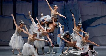 A large group of ballet dancers, seven women and four men, all on snowy stage. The women are in matching dresses and headwear that give off a snowy and icy look, and the men are in trousers and head dresses that look like wolf pelts. The dancers are all at different levels, some stood, some squat, some being lifted, and all pointing towards a male dancer on the floor who is dressed like a prince