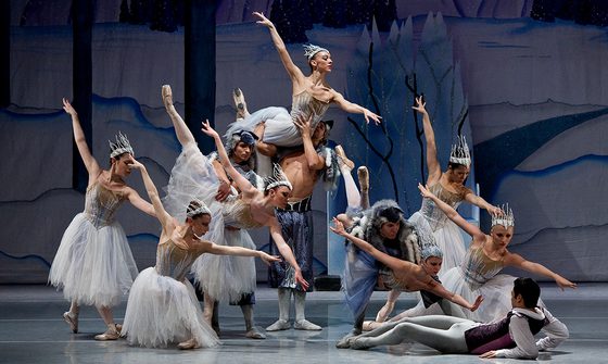 A large group of ballet dancers, seven women and four men, all on snowy stage. The women are in matching dresses and headwear that give off a snowy and icy look, and the men are in trousers and head dresses that look like wolf pelts. The dancers are all at different levels, some stood, some squat, some being lifted, and all pointing towards a male dancer on the floor who is dressed like a prince