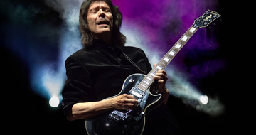 An older man with shoulder length brown hair closes his eyes as he plays on his black guitar