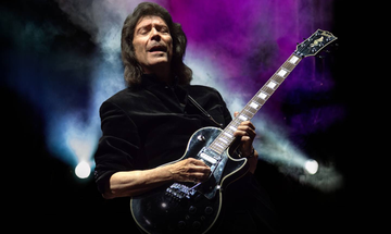An older man with shoulder length brown hair closes his eyes as he plays on his black guitar