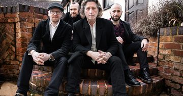 Four older white men in black suits sitting on brick steps outside a house.