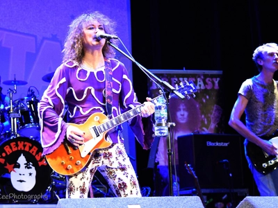Lit by blue light, a man with long curly hair wearing a purple flames top, sings into a microphone while playing a guitar, behind him is the rest of the band