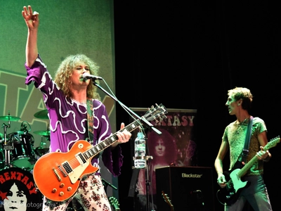 Lit by green light, a man with long curly hair wearing a purple flames top, sings into a microphone, holding his guitar with one hand and the other hand up in the air, the rest of his band are behind him