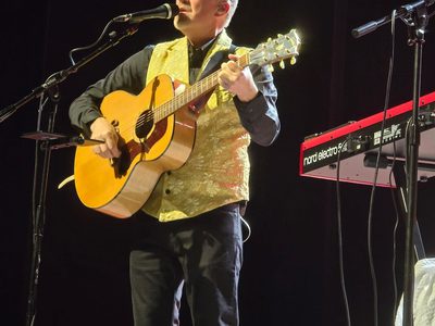 A man in an all black outfit with a yellow waistcoat stands playing his acoustic guitar while singing into a microphone
