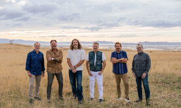 All six members of Toast stand in casual clothing in the middle of a field. The grass has dried so it's yellow and there's a slight orange tint to the sky amongst the clouds.