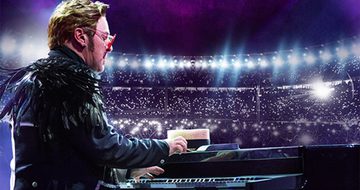 An Elton John impersonator plays guitar in front of an audience, wearing a black jacket and red glasses.