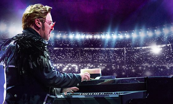 An Elton John impersonator plays guitar in front of an audience, wearing a black jacket and red glasses.
