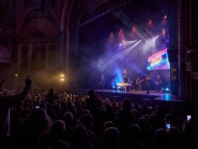Audience view of a live band performing on stage with a piano, while a large screen displays the words “Saturday Night's Alright.”