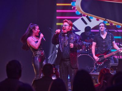 Audience view of an Elton John impersonator singing beside a female vocalist, with a guitarist and drummer in the background. The band members are all wearing black.