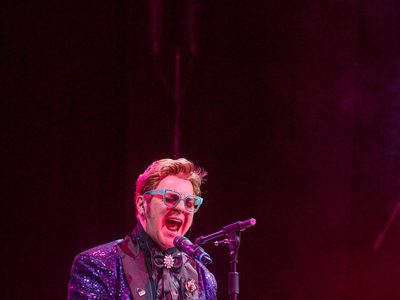 An Elton John impersonator performs on stage, wearing a sparkly suit jacket and blue glasses while playing the piano and singing into a microphone.