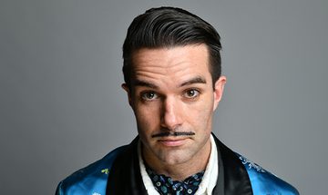 Troy Hawke, a man with a slicked-back hairstyle and thin mustache, wearing a blue and black patterned jacket, on a grey background
