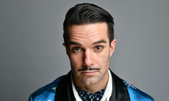 Troy Hawke, a man with a slicked-back hairstyle and thin mustache, wearing a blue and black patterned jacket, on a grey background