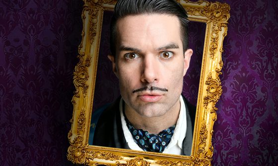 Troy Hawke, a white man with black hair and a marker pen pencil moustache, in a blue smoking jacket and cravat. His head is poking through a golden frame on a purple background
