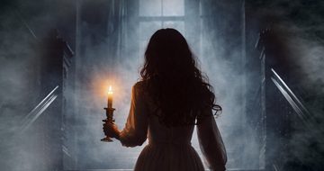 a lady walks up some fog covered stairs. She wears a long floaty white dress and holds a lit candle holder
