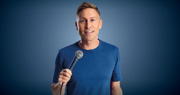 Russell Howard stands against a blue background, wearing a blue top and jeans and holding a microphone