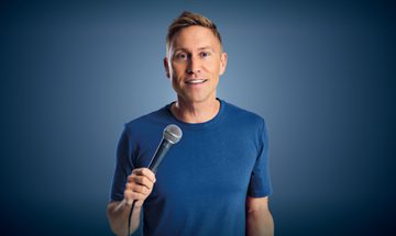 Russell Howard stands against a blue background, wearing a blue top and jeans and holding a microphone
