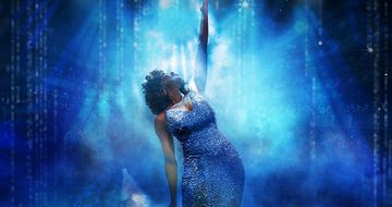 An image of a Whitney Houston impersonator in a sparkly blue dress holding a microphone with the other hand high in the arm that she is looking up at. The background is all blue