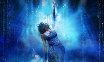 An image of a Whitney Houston impersonator in a sparkly blue dress holding a microphone with the other hand high in the arm that she is looking up at. The background is all blue