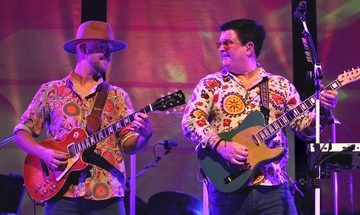 Two white men in colourful patterned shirts playing electric guitars on stage together