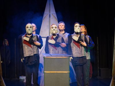 Three actors stand on a dimly blue-lit stage with fog, holding puppets positioned to appear as though the puppets have their hands on their chests.