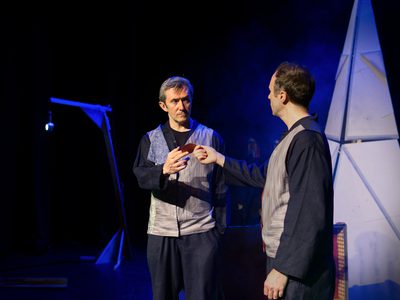 Two male actors stand on a dimly blue-lit stage, looking at each other with serious expressions. One, with his back to the camera, hands the other a red ticket.