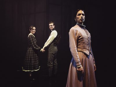 Three actors in period costumes on a dimly lit stage; a woman in pink stands in the foreground, while a man and woman hold hands in the background.