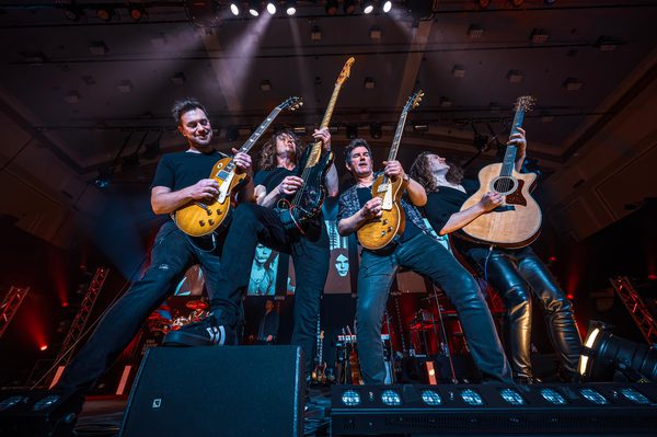 Four men perform on stage with guitars, with one musician resting a foot on a speaker.