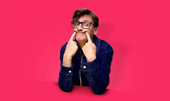 Ed Byrne, a white man with short quiffed brown hair and glasses, poking both of his index fingers into the corners of his mouth making an odd smile
