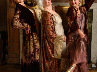 Members of Fascinating Aïda smiling together, leaning by a fireplace with photo frames above.