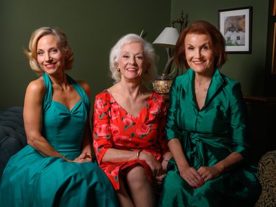 Members of Fascinating Aïda smiling together, dressed in elegant teal and red outfits, seated in a stylish, warmly lit room.