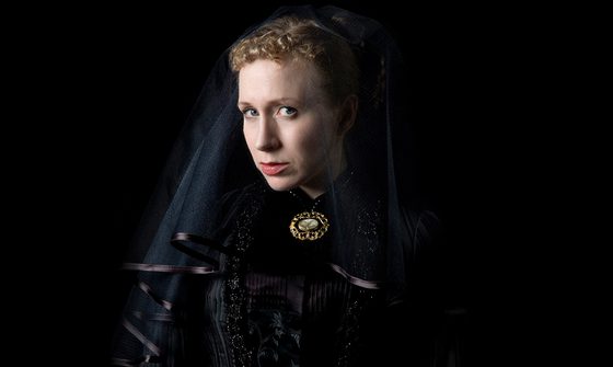 A white woman with curly blonde hair wearing a Victorian era black dress with a large gold pendant necklace and a black veil covering her face