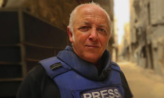 Jeremy Bowen an older balding white man with thinning white hair wearing a blue protective vest with 'PRESS' in white, over black shirt. He is standing in a war-damaged street.