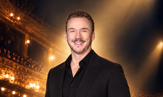 Russell Watson smiling and facing forwards, wearing a black suit with a busy auditorium and stage lights behind him.