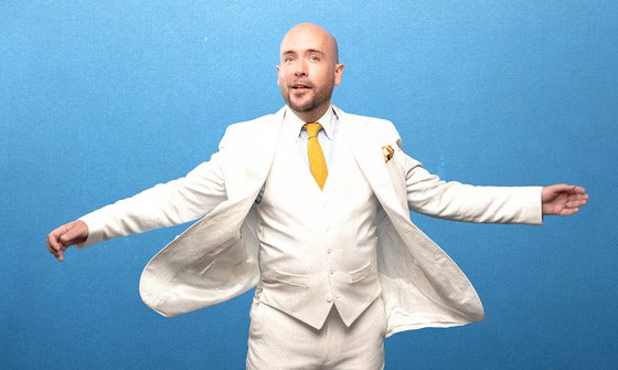 Tom Allen, a bald white man wearing a white blazer, waistcoat, shirt, and trousers, with a yellow tie stood with his arms out and blazer flying behind him on a blue background