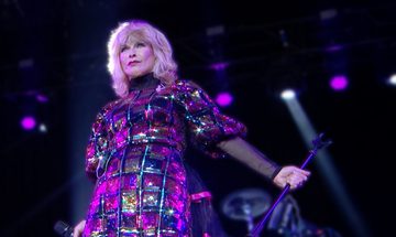 Toyah standing on stage in front of a set of drums. She is holding a microphone.