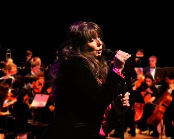 Mandy Watson, has long brown hair with a fringe, she's wearing a black blazer and sings into a handheld microphone in front of the orchestra.