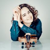 Alena Baeva leaning on a table with her arms raised towards her face, smiling at the camera with a violin on the table in front of her