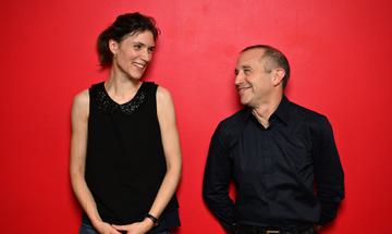 Chloé van Soeterstède and Mark Wigglesworth both wearing black and smiling at each other, standing against a bright red wall
