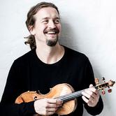 Christian Tetzlaff holding a violin, wearing a black long sleeve top, looking away from the camera and smiling against a plain white background