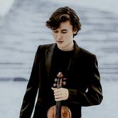 Daniel Lozakovich wearing a black top and black suit jacket, looking down and holding a violin