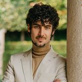Federico Colli wearing a light coloured jacket, leaning against a neutral wall outdoors, smiling at the camera