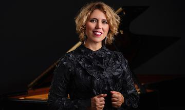 Gabriela Montero stood in front of a piano on a dark background, wearing a black dress and smiling at the camera
