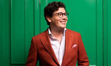 Martin James Bartlett stood against a green door, wearing a red blazer and smiling away from the camera