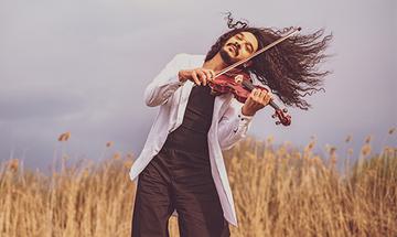 Nemanja Radulovic standing outside in long grass, playing a violin with his long hair blowing in the wind
