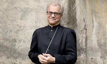 Osmo Vanska wearing a black long sleeve top and black rimmed glasses, holding a baton and smiling at the camera, standing in front of a neutral colour wall