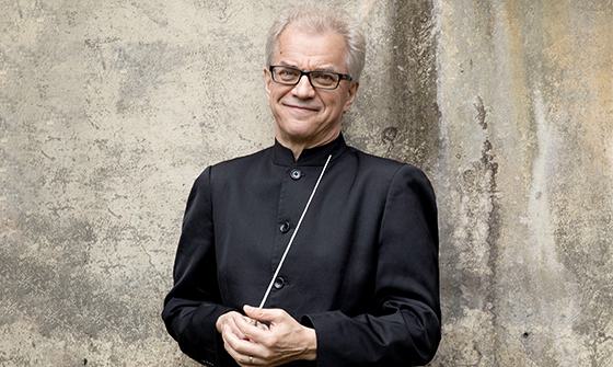 Osmo Vanska wearing a black long sleeve top and black rimmed glasses, holding a baton and smiling at the camera, standing in front of a neutral colour wall