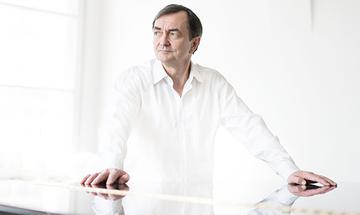 Pierre-Laurent Aimard wearing a white shirt, with both hands resting on the top of a piano in a bright white room, looking away from the camera