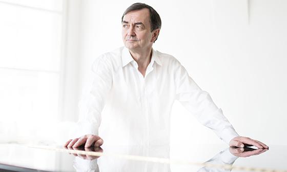 Pierre-Laurent Aimard wearing a white shirt, with both hands resting on the top of a piano in a bright white room, looking away from the camera